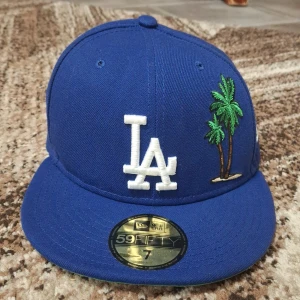 Blå LA keps från New Era 59FIFTY - Säljer en blå LA keps från New Era 59FIFTY med broderad vit LA-logga och palmer på framsidan. Kepsen har platt skärm och broderad MLB-logga bak. Storlek 7=55.8cm