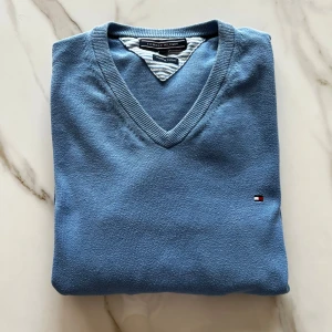 Blå v-ringad tröja från Tommy Hilfiger - Säljer en stilren blå v-ringad tröja från Tommy Hilfiger. Tröjan är i bomull och har en liten broderad logga på bröstet. Perfekt för dig som gillar klassisk och enkel stil. Långa ärmar och ribbade muddar.