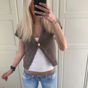 Brun stickad väst med knapp - Säljer en brun stickad väst med kort ärm och v-ringning. Västen har en enkel knapp framtill och är croppad i modellen. Perfekt att styla över en t-shirt eller linne.