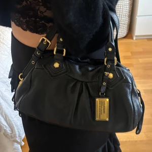 Svart Q Classic Baby Groove väska! - Snygg svart handväska i bra skick från Marc by Marc Jacobs i äkta läder med gulddetaljer och dubbla handtag (det långa finns med). Finns lite slitage på fötterna på undersidan, men inget tydligt alls. Små slitningar finns på några gulddetaljer, men inget stort. Välbevarad vintage modell! Perfekt för att ge en stilren touch till din outfit. Skriv privat för fler bilder. Mått 34x18x12 (undersida). Kan hämtas hemma hos mig i Nacka eller skickas men kan tyvärr inte mötas🤍