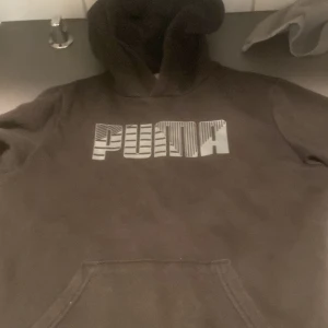 Svart hoodie från Puma - Säljer en svart hoodie från Puma med stor logga framtill i vitt och grått. Tröjan har huva och en klassisk magficka. Perfekt för dig som gillar sportig och avslappnad stil.