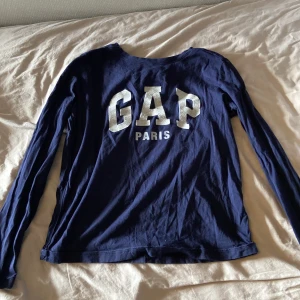 Mörkblå långärmad tröja från GAP - Säljer en mörkblå långärmad tröja från GAP med trycket 'GAP PARIS' i vitt framtill. Tröjan har en klassisk passform och är gjord i mjuk bomull, perfekt för en avslappnad stil.