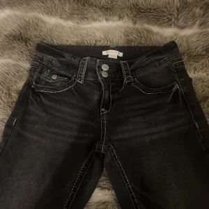 Svarta jeansbyxor från Gina Tricot - Säljer ett par svarta jeansbyxor från Gina Tricot med vita kontrastsömmar och klassisk femficksdesign. Byxorna har normal passform och stängs med dragkedja och knapp framtill. Perfekta till vardags för en stilren look.