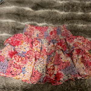 Blommig kortkjol från Kappahl - Supersöt kortkjol från Kappahl med blommigt mönster i rosa, rött, lila och gult. Kjolen har volangdetaljer och smock i midjan för en bekväm passform. Perfekt för vår och sommar!