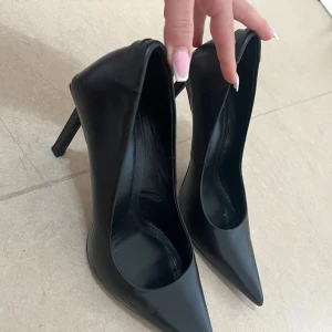 Svarta pumps från Zara - Klassiska svarta pumps från Zara med spetsig tå och hög klack. Skorna är tillverkade i syntetmaterial och har en elegant, stilren design som passar perfekt till många olika outfits.