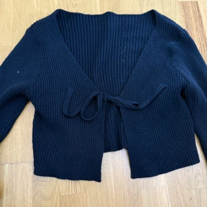 Svart ribbad kofta med knyt - Säljer en svart ribbad kofta med croppad passform och knyt framtill. Koftan har lång ärm och djup v-ringning, perfekt att slänga över en topp eller klänning.