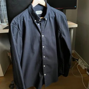 Mörkblå Oxford-skjorta från East West - Säljer en stilren mörkblå Oxford-skjorta från Brothers-East West, storlek L (passar även M). Endast använd en gång och i mycket fint skick. Det finns ett litet lagat märke till vänster som knappt syns. Perfekt för både festliga tillfällen och en avslappnad, casual stil. 