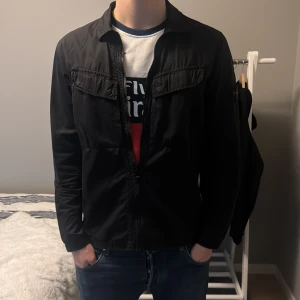 Stone Island overshirt  - Svart overshirt från Stone Island med klassisk krage och dragkedja framtill. Jackan har två bröstfickor och den ikoniska Stone Island-loggan på vänster ärm. Perfekt för en stilren och avslappnad look. Modellen är 190 och 80kg. 