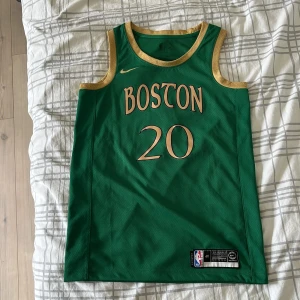 Boston Celtics baskettröja Hayward #20 - Grön baskettröja från Nike med guldiga detaljer och texten 'BOSTON' samt nummer 20 på framsidan. På baksidan står det 'HAYWARD' och nummer 20. Tröjan är ärmlös och tillverkad i Dri-Fit material för bra ventilation. NBA-logga och etikett nertill.