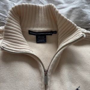Beige stickad kofta från Ralph Lauren Sport - Säljer en beige stickad kofta från Ralph Lauren Sport med dragkedja och kabelstickade detaljer längs ärmarna. Tröjan har hög krage och ribbade muddar. Klassisk broderad logga på bröstet. Står strl L men passar som en mindre M.