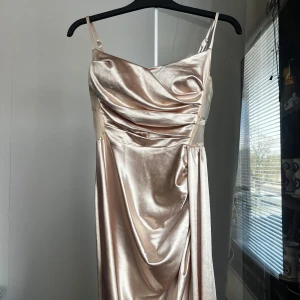Champagnefärgad långklänning med drapering - Säljer en elegant långklänning i skimrande champagnefärgat satin med smala axelband och draperad framsida. Baksidan har meshparti med broderade detaljer i guld. Perfekt för bal eller fest! (Använt en gång)