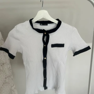 Vit ribbad blus med svarta detaljer från Zara - Säljer en vit ribbad blus från Zara med svarta volangdetaljer vid krage, ärmslut och knappslå. Blusen har korta ärmar, dekorativ ficka och guldknappar framtill. Perfekt för en stilren och trendig look.
