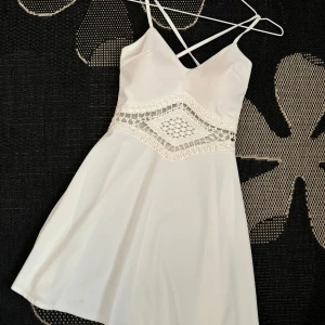 Vit klänning med spetsdetaljer och smala axelband - Säljer en vit klänning med smala axelband och v-ringning. Klänningen har fina virkade spetsdetaljer i midjan. Lapp med storlek saknas, passar XS. 