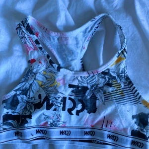 Blommig sport-bh från Stadium - Säljer en sport-bh från Stadium med blommigt mönster i svart, vitt och inslag av gult och rosa. Modellen har brottarrygg och elastiskt band nertill med WARP-logga. Perfekt för träning eller sportiga aktiviteter.