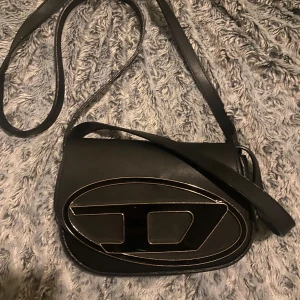 Svart axelväska med stort Dmärke - Säljer en svart axelväska från disel med ett stort D format metallmärke på framsidan. Väskan har justerbar axelrem och magnetlås. Insidan är fodrad i ljust tyg och har ett innerfack.