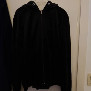 Svart hoodiejacka med dragkedja - Svart hoodiejacka med dragkedja framtill och huva. Jackan har en mjuk, sammetsliknande yta och långa ärmar. Perfekt för en avslappnad stil.