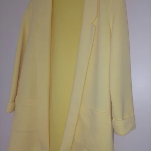 Gul kavaj från Amisu - Snygg ljusgul kavaj från Amisu med långa ärmar och två stora fickor framtill. Kavajen har en klassisk krage och är tillverkad i ett strukturerat tyg som ger en stilren look. Perfekt för att liva upp din outfit med lite färg.