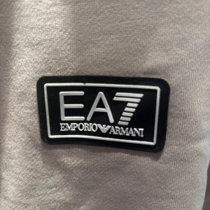 Beige sweatshirt från EA7 Emporio Armani - Säljer en beige sweatshirt från EA7 Emporio Armani med svartvit logga på bröstet och dekorativa band med logotyp längs ärmar och rygg. Tröjan har rund hals och långa ärmar, perfekt för en stilren och sportig look. Köpt för 1000kr, mitt pris:450kr. Kontakta gärna vid frågor. Skick: ny testad en gång.