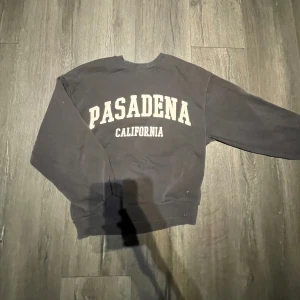 Svart sweatshirt med Pasadena-tryck - Cool svart sweatshirt med texten 'PASADENA CALIFORNIA' i vitt framtill. Klassisk rund hals och långa ärmar. Perfekt för en avslappnad stil.