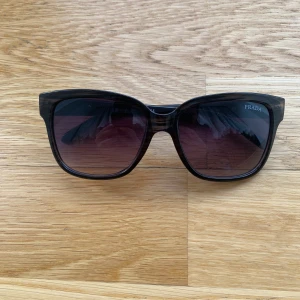 Svarta solglasögon från Prada - Snygga svarta solglasögon från Prada med fyrkantig form och mörka glas. Bågarna har diskreta gulddetaljer på sidorna där loggan syns. Har använts ett par gånger. Klassisk och stilren design som passar till många tillfällen.  Pris kan diskuteras.