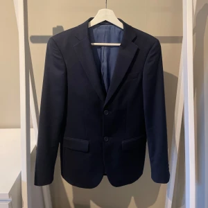 Mörkblå kavaj från Dressmann - Säljer en stilren mörkblå kavaj från Dressmann i slim fit-modell. Kavajen har två knappar framtill, fickor med lock och är helfodrad med ljusblå insida och röd. Säljer den pågrund av att den har blivit för liten, nypris: 1000, mitt pris 499.                   Tveka inte på att höra av dog vod frågor!