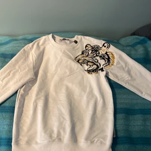 Vit sweatshirt med tigerbroderi från Philipp Plein  - Säljer en vit sweatshirt från Philipp Plein med ett stort broderat tigerhuvud i svart och guld på bröstet. Tröjan har rund halsringning och ribbade muddar. Perfekt för dig som vill sticka ut med en unik detalj.