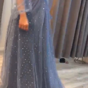 Blå glittrig långklänning med volangärmar - Säljer en blå långklänning med glittriga detaljer och transparenta volangärmar. Klänningen har v-ringning och markerad midja med knappar framtill. Perfekt för bal eller festliga tillfällen.