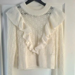 Vit stickad tröja med volangdetaljer - Säljer en vit stickad tröja med genombrutet mönster och stora volanger framtill och över axlarna. Tröjan har lång ärm och ribbade muddar vid hals, ärmslut och nederkant. Perfekt för dig som gillar romantisk stil!