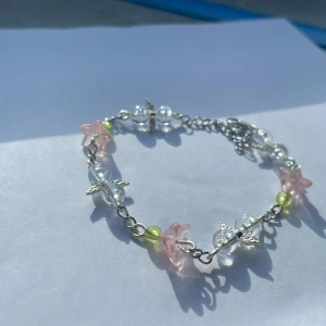 Handgjort armband med pärlor och blomdetaljer - Handgjort armband med genomskinliga, små gröna och ljusrosa blomformade pärlor🌸. Utsmyckat med små silverfärgade vingar. Justerbar längd med karbinlås. Inte äkta silver💕 Ge gärna prisförslag!