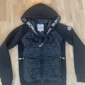 Svart quiltad hoodie från Moncler - Svart hoodie från Moncler med quiltad framsida och dragkedja. Stor huva med Moncler-logga längs kanten och två fickor med dragkedja. Klassisk Moncler-patch på ärmen. Perfekt för en sportig och stilren look.