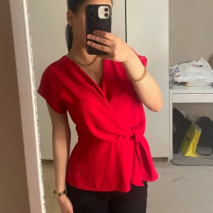 Röd omlottblus från Zara - Säljer en snygg röd omlottblus från Zara i storlek XS. Blusen har korta ärmar, knyt i midjan och en v-ringad halsringning. Perfekt för dig som vill ha en färgstark och stilren topp till garderoben.