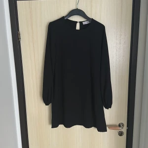 Svart klänning från Style State - En stilren svart klänning från Style State med rund halsringning och långa ärmar. Klänningen har en rak passform och en knappdetalj i nacken. Perfekt för dig som gillar enkel och klassisk stil.