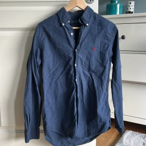 Mörkblå skjorta från Ralph Lauren - Klassisk mörkblå skjorta från Ralph Lauren med button-down krage och broderad röd logga på bröstet. Skjortan har långa ärmar och vita knappar framtill samt vid ärmsluten. Perfekt för en stilren look.