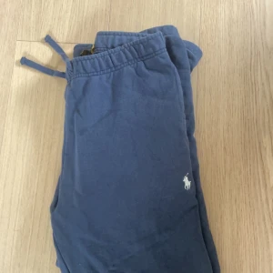 Blå mjukisbyxor från Polo Ralph Lauren - Mörkblå mjukisbyxor från Polo Ralph Lauren med dragsko i midjan och broderad vit logga på ena benet. Perfekta för chill dagar eller när du vill vara extra bekväm. Klassisk och enkel design.