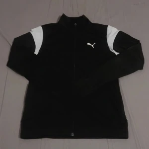 Svart träningsjacka från Puma - Säljer en svart träningsjacka från Puma med vita detaljer på axlarna och dragkedja framtill. Jackan har hög krage och lång ärm, perfekt för träning eller sportiga outfits. Puma-logga på bröstet.