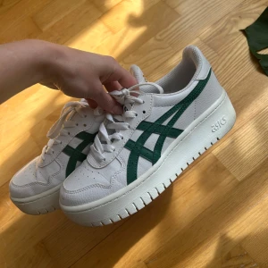 Vita sneakers från Asics med gröna detaljer - Säljer ett par fräscha vita Asics sneakers med gröna ränder på sidorna. Skorna har snörning, rund tå och en rejäl platt sula som ger extra höjd. 