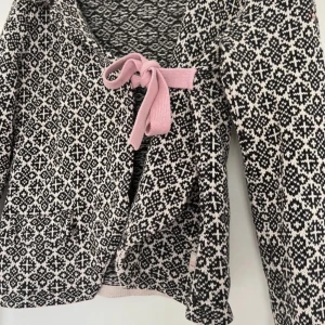 Svartvit stickad jacka med rosett från Odd Molly - Säljer en svartvit stickad jacka från Odd Molly med klassiskt mönster och en rosa rosett framtill. Jackan har lång ärm och är i mjukt material, perfekt för lager-på-lager. Snygg och unik detalj med broderad logga på ärmen.