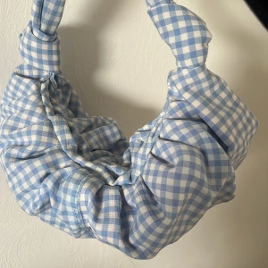 Blåvit rutig axelväska - Superfin axelväska i blåvitt rutigt tyg med knutdetaljer på handtaget. Väskan har en mjuk och puffig form som ger en trendig look. Perfekt för att addera lite färg och mönster till din outfit!