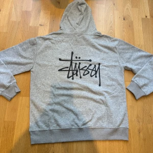 Grå hoodie från Stüssy - Säljer en grå hoodie från Stüssy med svart logga tryckt på bröstet och ryggen. Tröjan har huva med dragsko och ribbade muddar vid ärmslut och nederkant. Perfekt för en avslappnad streetwear-look.