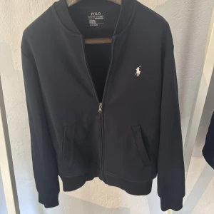 Marinblå zip-tröja från Polo Ralph Lauren - Säljer en marinblå zip-jacka från Polo Ralph Lauren med klassisk broderad logga på bröstet. Jackan har två fickor framtill, ribbade muddar och krage. Perfekt för en stilren och enkel look.