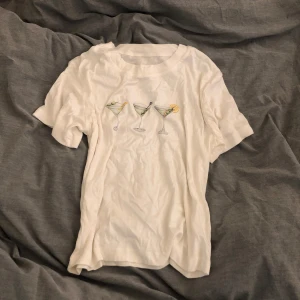 Vit t-shirt med print  - Vit t-shirt med tryck av tre martiniglas med oliver och citron på bröstet. Klassisk rund halsringning och korta ärmar. Perfekt för dig som gillar roliga och unika motiv på dina kläder. I bra skick 💖
