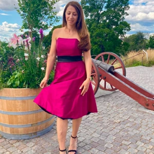 Axelbandslös ceriserosa klänning med svart midjeband - Säljer en axelbandslös ceriserosa klänning med svart band i midjan. Klänningen har en utställd kjol och slutar ovanför knäna. Perfekt för sommarens tillställningar!