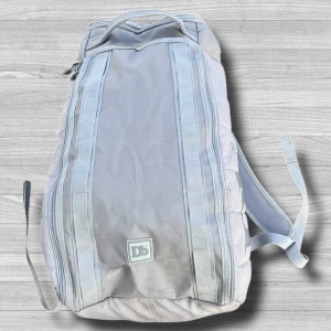 Douchebag - Säljer en stilren ljusgrå ryggsäck från Db med minimalistisk design och diskret logga framtill. Väskan har flera dragkedjefack och justerbara axelremmar för bekväm passform. Perfekt för dig som vill ha en snygg och praktisk accessoar till vardagen.