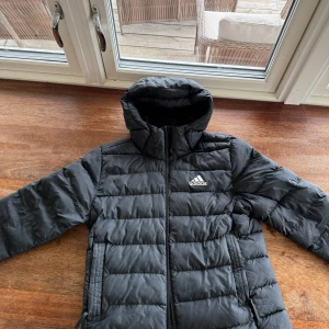 Svart dunjacka från Adidas med huva - Säljer en svart dunjacka från Adidas med huva och dragkedja. Jackan har en klassisk pufferstil med horisontella sömmar och två fickor framtill. Perfekt för kyliga dagar och enkel att matcha med det mesta.