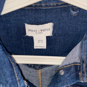 Jeansklänning från Holly & Whyte - Snygg jeansklänning från Holly & Whyte med långa ärmar, guldknappar framtill och två bröstfickor. Klassisk krage och markerad midja ger en tidlös look. Perfekt för dig som gillar denim och vill ha en stilren klänning.