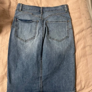 Jeanskjol med klassisk femficksdesign - Säljer en blå jeanskjol med normal passform och klassisk femficksdesign. Kjolen har bälteshällor, knapp och dragkedja framtill samt raka linjer för en stilren look. Perfekt för dig som gillar enkel denimstil. Storlek XS