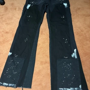 Svarta flared jeans med vita färg detaljer  - Unika svarta jeans med breda ben och vita färgstänk samt råa, ofållade kanter längst ner. Byxorna har paneler i olika svarta nyanser och en cool, konstnärlig look. Perfekta för dig som vill sticka ut med din stil.