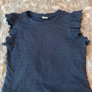 Mörkblå topp med volangärmar - Säljer en mörkblå topp med korta volangärmar och rund halsringning. Toppen har ett diskret prickigt strukturmönster i tyget och är tillverkad i ett mjukt material. Perfekt för en avslappnad men ändå söt look. Storlek 134-140 i barnstorlek