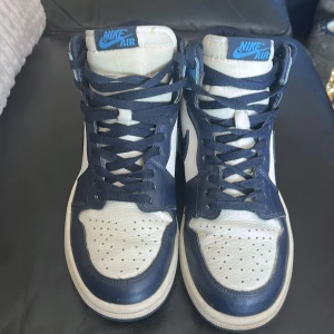 Nike Air Jordan 1 High blå/vit - Säljer ett par Nike Air Jordan 1 High i blått, vitt och marinblått skinn. Klassisk siluett med högt skaft, perforerad tå och blå Nike Air-logga på plösen. Snygga kontraster och ikonisk Jordan Wings-detalj på sidan. Perfekta för dig som gillar streetstyle.