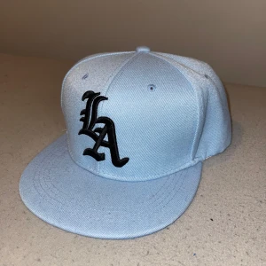 Ljusblå LA keps med broderad logga - Snygg ljusblå snapback-keps med svart broderad LA-logga framtill. Kepsen har platt skärm och justerbar plastrem baktill för perfekt passform. Tillverkad i bomullstyg som känns både mjukt och slitstarkt. Perfekt accessoar för en cool streetstyle.
